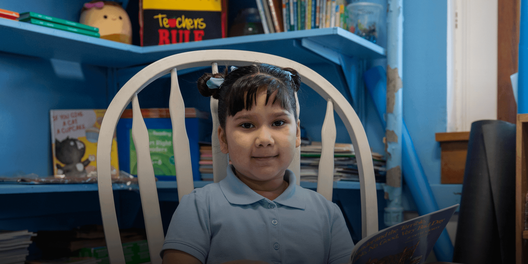Young student in a light blue uniform shirt reading a book in a classroom reading corner.