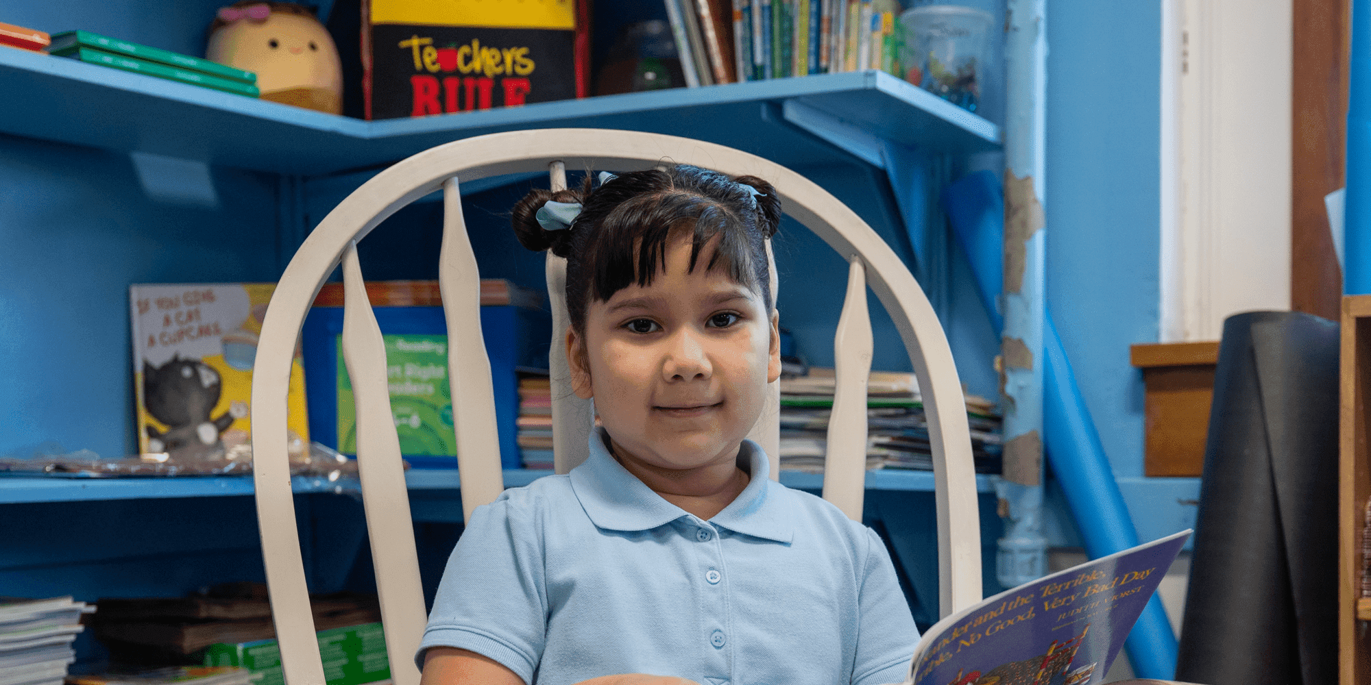 Young student in a light blue uniform shirt reading a book in a classroom reading corner.